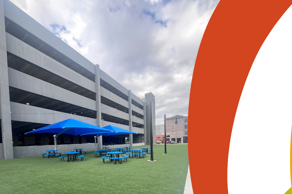 Photo of The Fringe, blue picnic tables under a blue umbrella on green turf in front of the parking garage in Bowling Green, Kentucky